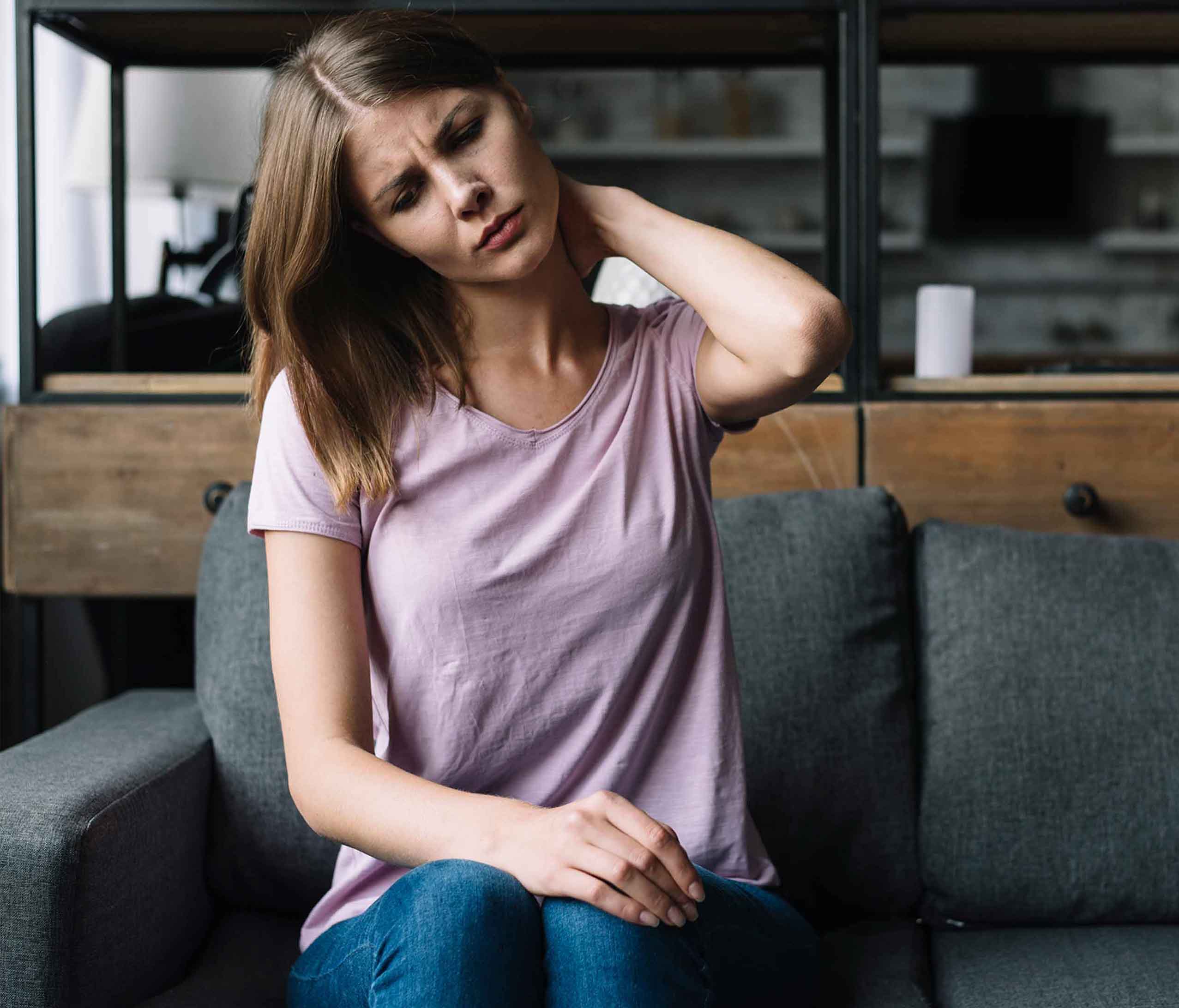 Mujer joven con malestares en la zona del cuello; esta se toca el área del cuello con una expresión de dolor. Ella se encuentra sentada en el sillón de su sala.